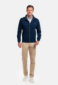 Navy zip-up jacket with stand collar, ribbed cuffs, and contrasting trim; worn over a patterned shirt, paired with beige pants and white sneakers.