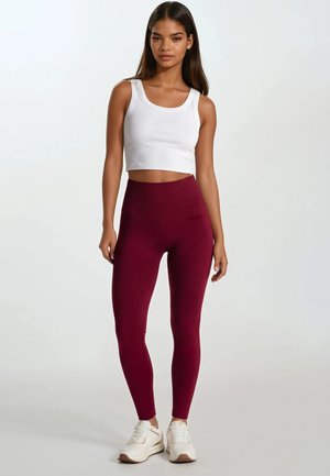 Young woman standing wearing white cropped tank top, high-waisted burgundy leggings, and white sneakers on light background.