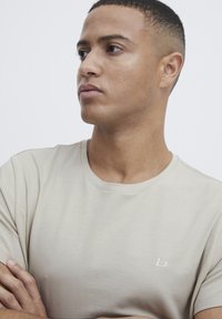 Beige crew-neck t-skjorte laget av myk bomull, med en subtil brodert logo på brystet og korte ermer. Glatt tekstur.