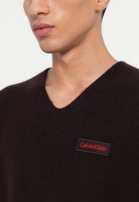 Σκούρο μωβ πουλόβερ V με μαλακό υλικό, διαθέτοντας ορθογώνια κόκκινη ετικέτα λογότυπου με "Calvin Klein" μπροστά.