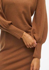 Pull marron avec des poignets côtelés et des manches bouffantes. La texture douce est visible, mettant en avant un design ajusté qui épouse le corps.