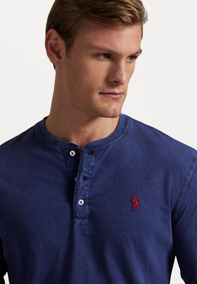 Μακρυμάνικη μπλούζα henley σε ναυτικό μπλε χρώμα, φτιαγμένη από μαλακό ύφασμα, με κουμπιά στον λαιμό και κόκκινο κεντητό λογότυπο στην αριστερή πλευρά του στήθους. Απαλή επιφάνεια.