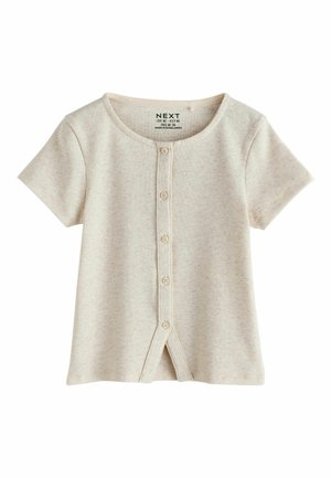 Mladé dievča stojí sebavedomo s rukami v bokoch, oblečené v krátky béžový top na gombíky, široké hnedé nohavice a ružovo-biele tenisky.