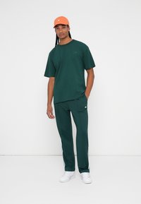 Mørkegrønn bomull T-skjorte og matchende joggebukser med hvite sidestriper. Oransje caps med logo. Hvite sneakers. Minimalistisk design.