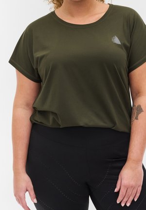 Mujer con camiseta de manga corta verde oscuro y leggings negros, mostrando tatuaje en el antebrazo izquierdo y cabello rizado parcialmente visible.