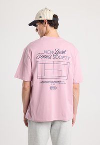 Pink cotton t-shirt featuring a printed tennis court design and text on the back. Paired with grey pants and a striped cap.