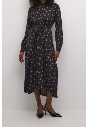 Woman wearing a black midi dress with long sleeves and brown polka dots, tied waist, button front, paired with black pointed heels.