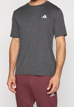 Homme portant un t-shirt gris foncé à manches courtes et un pantalon bordeaux, tous deux avec des logos Adidas blancs sur la poitrine et la cuisse.