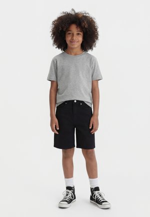 Enfant aux cheveux bouclés portant un t-shirt gris, un short noir, des chaussettes blanches et des baskets montantes noires, debout sur un fond blanc.