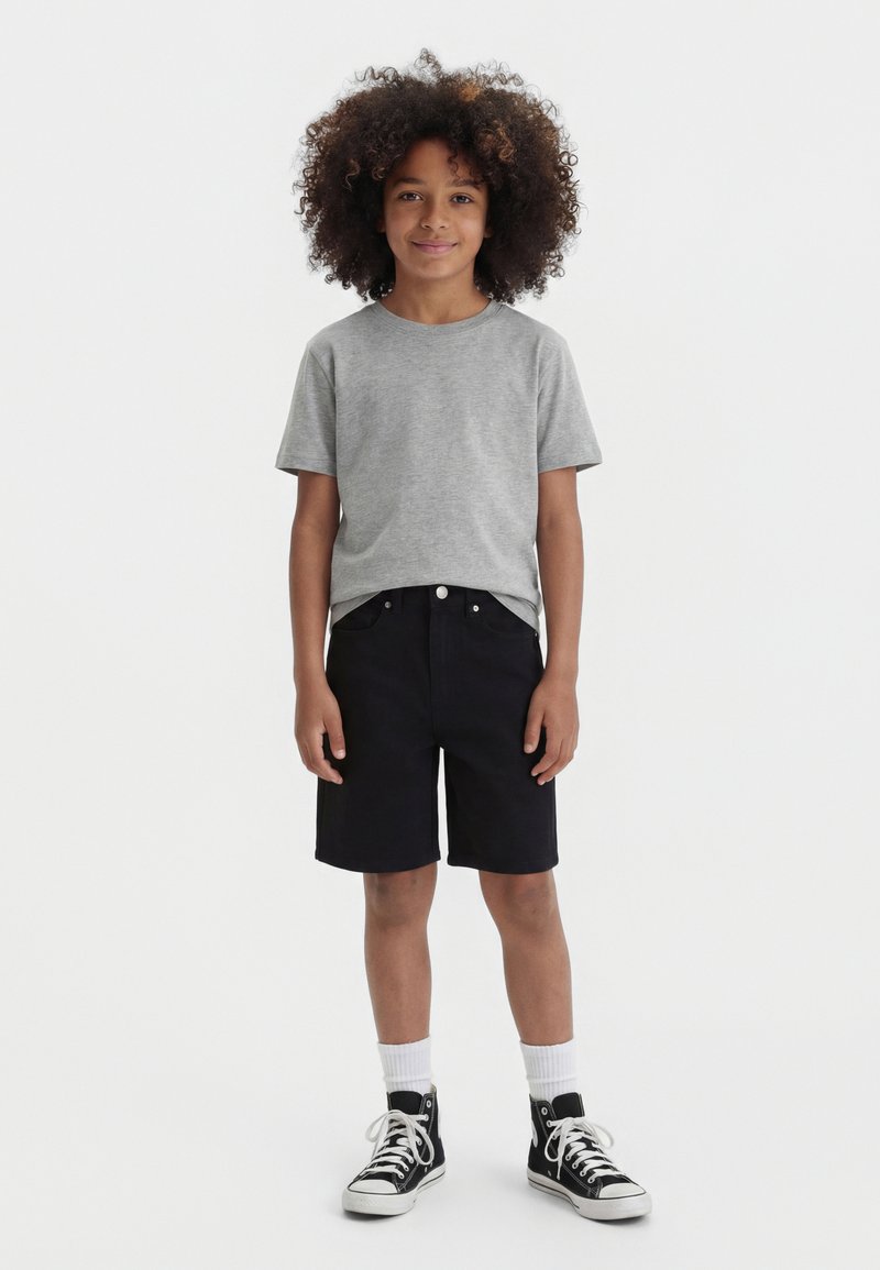 Enfant aux cheveux bouclés portant un t-shirt gris, un short noir, des chaussettes blanches et des baskets montantes noires, debout sur un fond blanc.