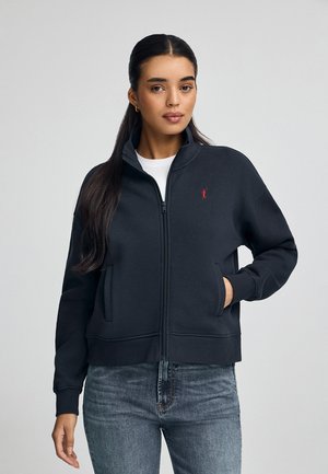 Sudadera con cremallera de color gris oscuro, con un alto cuello, dos bolsillos y un pequeño logo rojo en el lado izquierdo del pecho. Material de peso medio, ajuste casual.