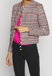 Flerfarvet tweedjakke uden krave, med frynse detaljer og lange ærmer, kombineret med en lyserød top og sorte jeans.