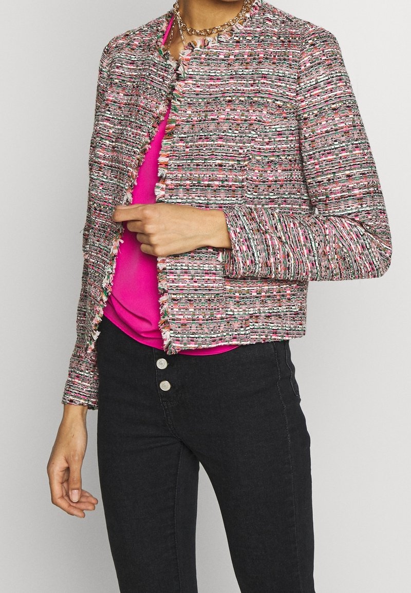 Kolorowa, tweedowa kurtka bez kołnierza z detalami w postaci frędzli i długimi rękawami, połączona z różową bluzką i czarnymi dżinsami.