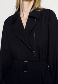 Chaqueta negra de tela suave, con cuello con muesca, cierre diagonal de cremallera y un cinturón ceñido con una hebilla de metal.