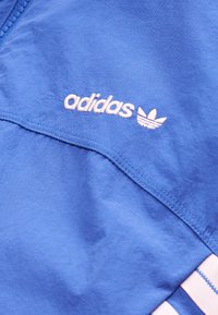 Μπλε ρούχο Adidas με λευκό κεντητό λογότυπο και τρεις λευκές ρίγες κατά μήκος της άκρης, που δείχνει την υφή του υφάσματος και τις λεπτομέρειες των ραφών.