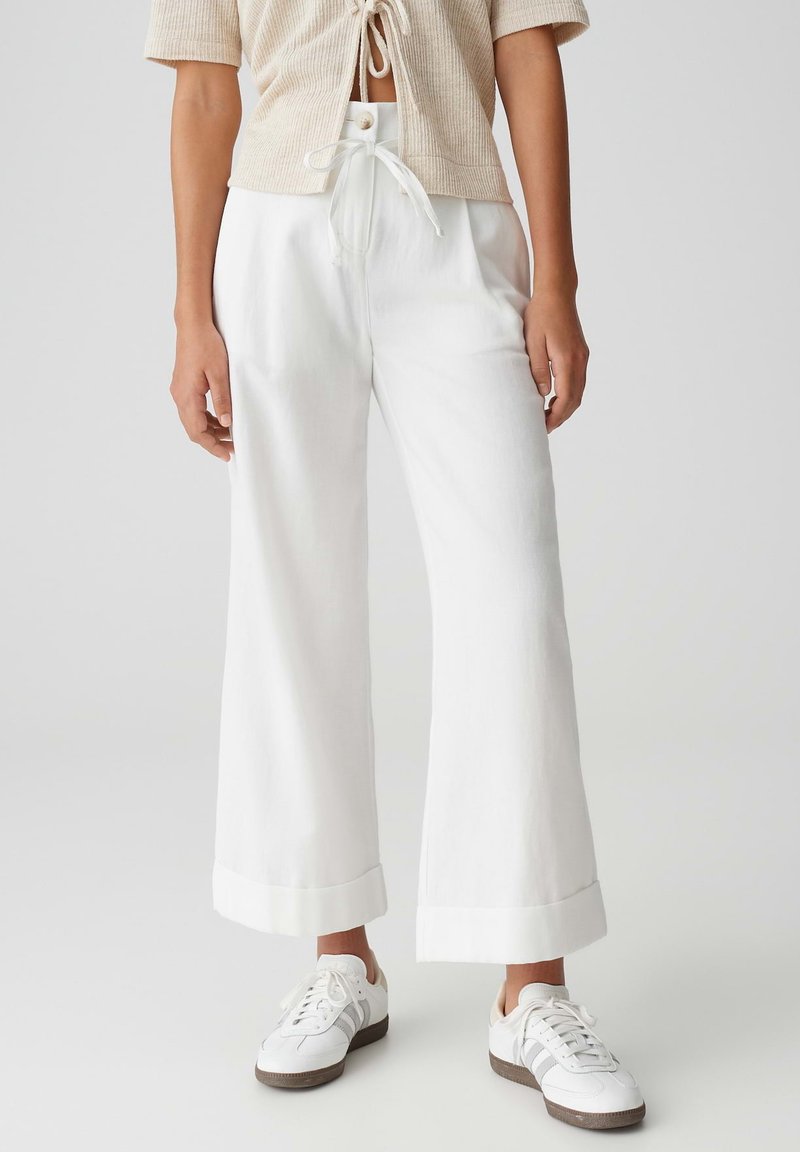 White wide-leg trousers made of lightweight fabric, featuring a high waist, pleats, and rolled hems. Paired with white sneakers.