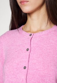 Vaaleanpunainen neulecardigan, jossa on pyöreä pääntie, etupuolella tummat napit ja pehmeä, teksturoitu pinta.