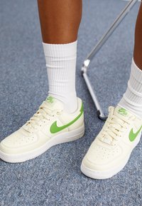 Bijele Nike Air Force 1 tenisice s zelenim detaljima, s glatkim kožnim gornjištem, perforacijama i bijelim teksturiranim potplatom.