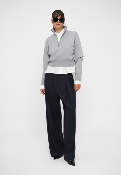 Pulover cu fermoar de culoare gri deschis, cu un tiv alb în straturi, asortat cu pantaloni largi de culoare bleumarin închis și pantofi negri cu vârf ascuțit. Ochelari de soare negri purtați.
