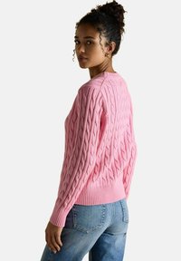 Kobieta z kręconymi włosami, ubrana w różowy sweter warkoczowy i niebieskie dżinsy, spoglądająca przez ramię na białym tle.