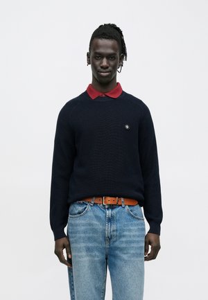 Homme à la peau foncée et aux cheveux tressés portant un pull bleu marine par-dessus un polo rouge, un jean bleu clair et une ceinture marron, debout devant un fond uni.
