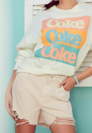 Cremefarvet sweatshirt med farverig "Coke" tekst i orange og blå, parret med lyse beige slidte shorts med frynset kant.