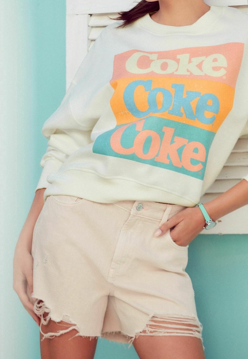 Cream sweatshirt with colorful "Coke" text in orange and blue, paired with light beige distressed shorts featuring frayed hem.