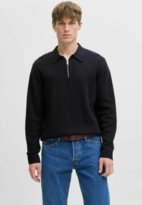 Jauns vīrietis, ģērbs ar melnu garu piedurkņu zipa džemperi ar apkakli un ziliem džinsiem, stāv pret vienkāršu gaišu fona.