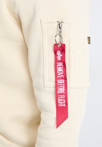 Sweatshirt beige avec une poche zippée, fermeture éclair en métal, et une étiquette en tissu rouge portant le texte "RETIRER AVANT LE VOL" attachée à la tirette de la fermeture.