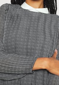 Texturerad grå blus med ett rutmönster, har en vit krage och ribbade lång ärmar. Tyget verkar mjukt och något stretchigt.