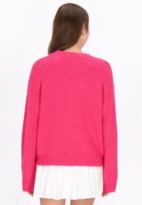 Pulover roz pufos, cu guler înalt și mâneci lungi, având o margine cu dungi. Modelul poartă o fustă albă plisată dedesubt.