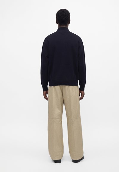 Homme vu de dos portant un haut noir à manches longues et un pantalon large beige, debout devant un fond blanc uni.