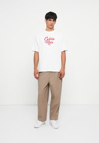 Camiseta blanca con logo rojo de "Calvin Klein", pantalones beige holgados, zapatillas blancas y cabello corto negro contra un fondo liso.