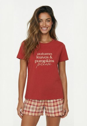 Kvinde smiler iført en rød T-shirt med teksten "autumn leaves & pumpkins please" og røde ternede shorts mod en ensfarvet hvid baggrund.