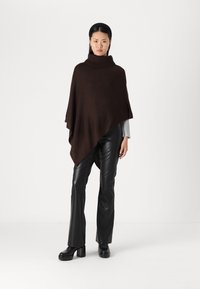 Brun strikket poncho med høy krage, båret over en lys grå topp og svarte skinnbukser, parret med chunky svarte platåsko.