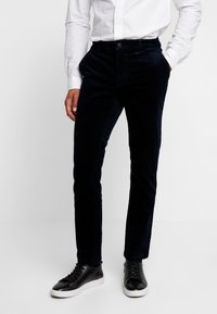 Tmavě modré kordové kalhoty s úzkým střihem, se zapínáním na knoflík a bočními kapsami, doplněné černými teniskami. Hladká textura.