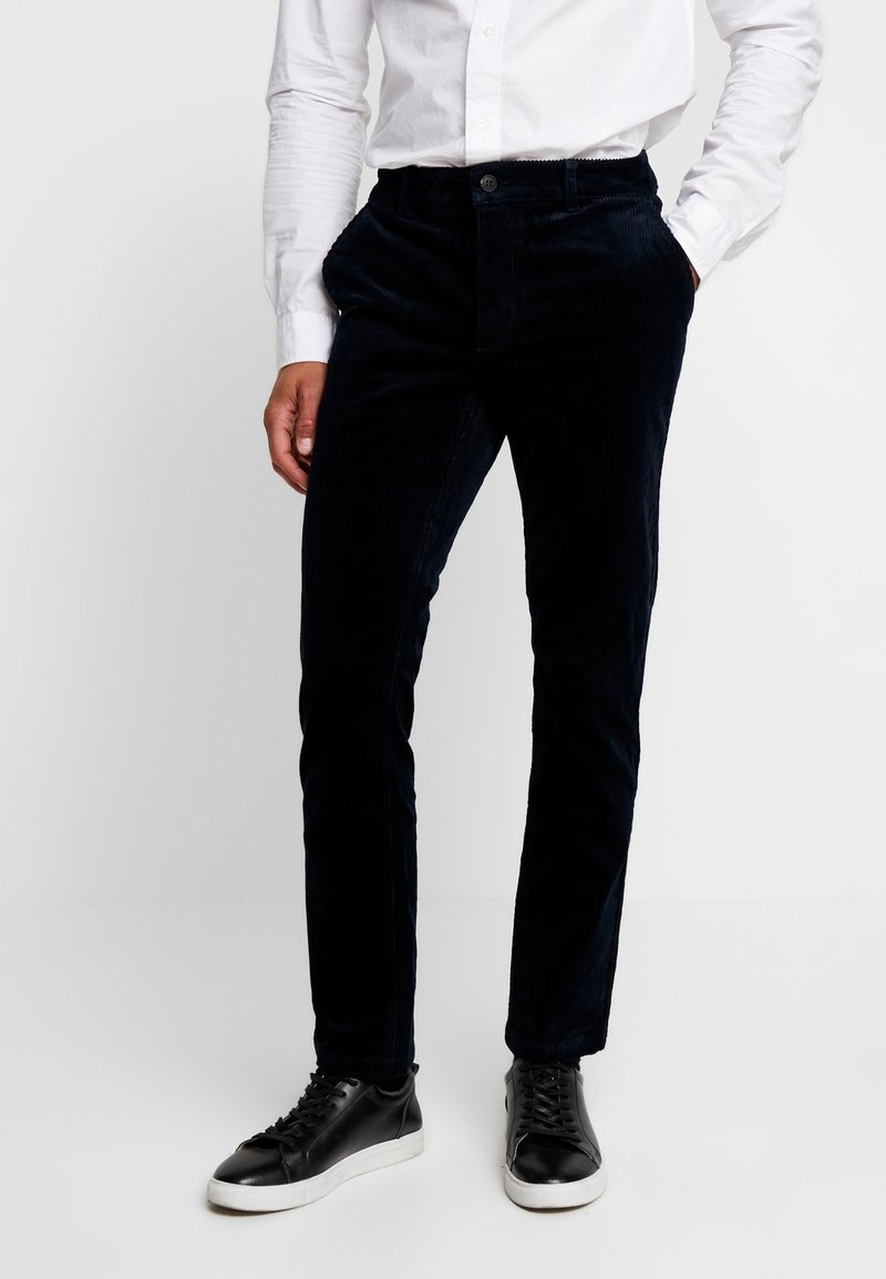 Tmavě modré kordové kalhoty s úzkým střihem, se zapínáním na knoflík a bočními kapsami, doplněné černými teniskami. Hladká textura.