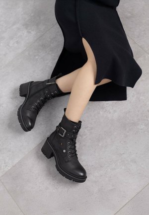 Botas de tobillo de cuero negro con un tacón robusto, parte delantera con cordones, cremallera lateral y una hebilla decorativa. Parte superior texturizada y suela resistente.