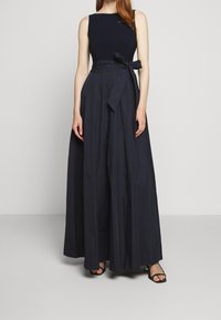 Kvinna i en ärmlös svart aftonklänning med hög midja, knytning och vid kjol, kombinerad med svarta öppna klackar.