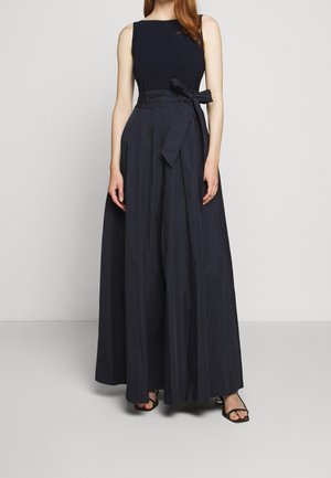 Femme portant une robe de soirée noire sans manches avec une taille haute et une jupe évasée, associée à des escarpins noirs ouverts.