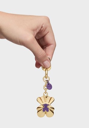 Mano sosteniendo un llavero dorado y morado con un pequeño recorte en forma de oso y una etiqueta redonda con la inscripción "TOUS".