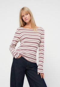 T-shirt à manches longues avec un col bateau, en tissu blanc avec des rayures horizontales bordeaux. La texture semble douce, associé à un pantalon en denim foncé ample.
