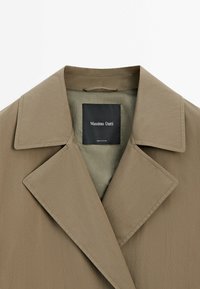 Hellbrauner Trenchcoat mit einem großen, eingeschnittenen Revers und glatter Textur. Innen befindet sich ein kontrastierendes hellgrünes Futter mit einem Markenetikett.