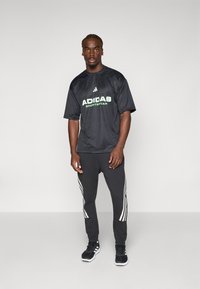 Zwart sportT-shirt met groen "ADIDAS Sportswear" logo, gecombineerd met zwarte broek met witte drie-strepen accenten. Gedragen met zwarte sneakers.