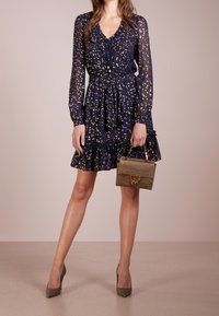 Marinblå klänning med gyllene prickar, volangnederdel, knytenacke, långa ärmar. Modellen håller en liten olivgrön handväska med ett gyllene spänne.