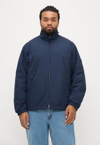 Geacă căptușită de culoare albastru marin, cu guler înalt, fermoar frontal și manșete elastice. Țesătură ușoară cu textură netedă și buzunare laterale.