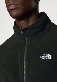 Tummanvihreä fleece-takki, jossa on korkea kaulus, vetoketjusuljenta ja valkoinen logo vasemmalla puolella. Pehmeä pinta ja istuva muotoilu.