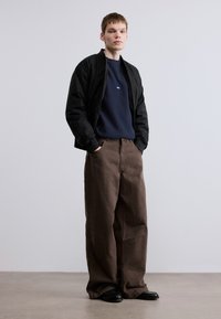 Μαύρο μπουφάν bomber πάνω από μπλε σκούρα μπλούζα με μικρό λογότυπο, σε συνδυασμό με φαρδιά καφέ παντελόνια και μαύρα παπούτσια, στέκεται μπροστά σε ουδέτερο φόντο.