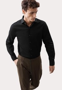 Camicia nera a maniche lunghe con colletto classico, abbinata a pantaloni marroni a pieghe con cordoncino. Tessuto dalla texture liscia.