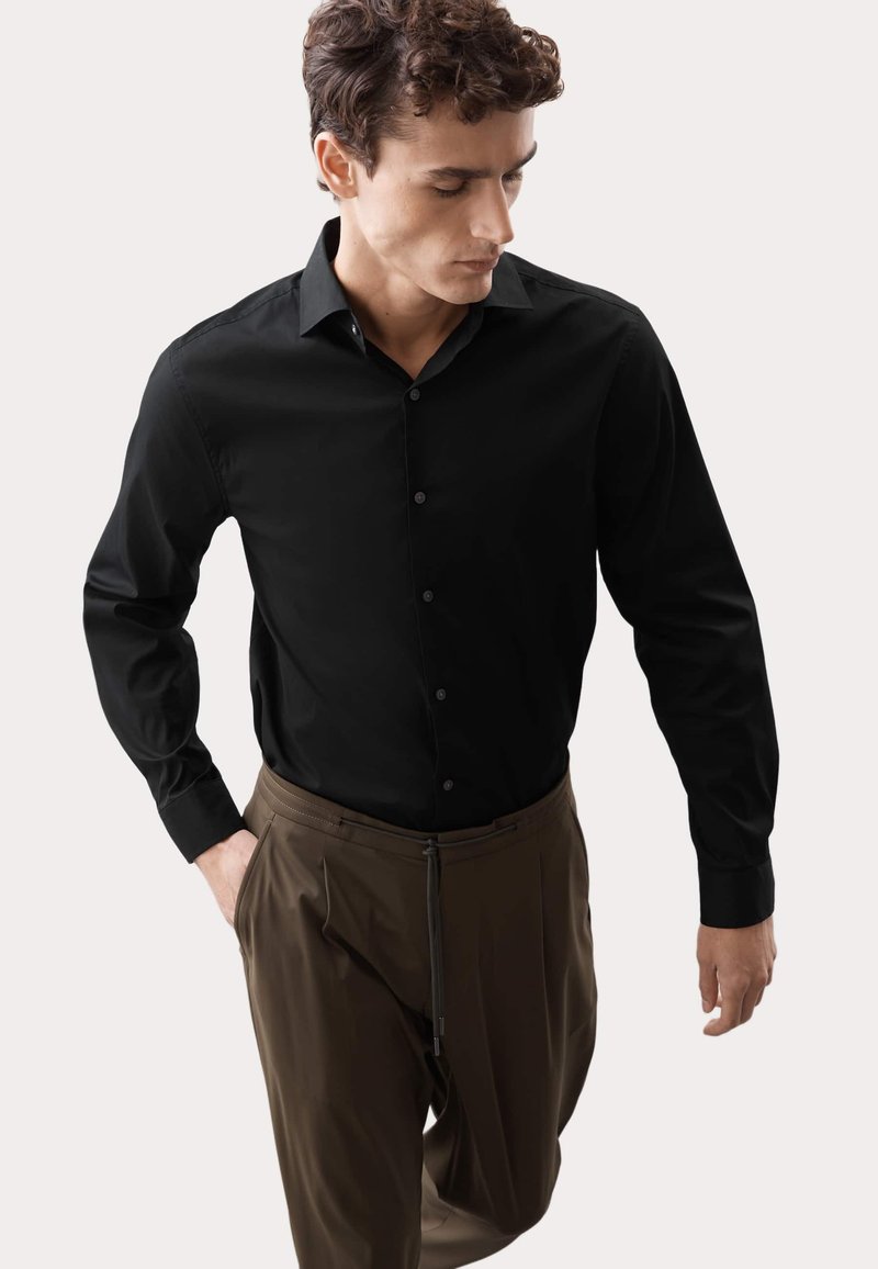 Camicia nera a maniche lunghe con colletto classico, abbinata a pantaloni marroni a pieghe con cordoncino. Tessuto dalla texture liscia.